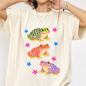 May include: Ivory-coloured t-shirt featuring three colourful frog illustrations. The frogs are green, red, and purple, each with unique patterns and surrounded by pink and blue stars. The shirt has a relaxed fit.