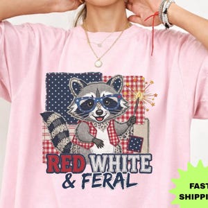 May include: A light pink t-shirt with a cartoon raccoon wearing glasses and holding a sparkler. The raccoon is set against a patchwork background with the text "RED WHITE & FERAL".
