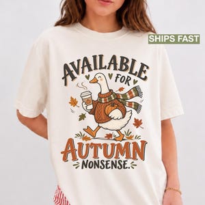 May include: An off-white t-shirt featuring a graphic of a goose wearing a brown sweater and a striped scarf, holding a coffee cup and a pumpkin. The text reads "AVAILABLE FOR AUTUMN NONSENSE" surrounded by falling autumn leaves in orange and brown tones.