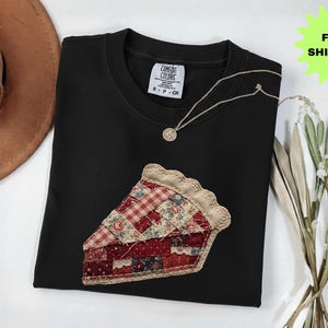 May include: A black crew neck sweatshirt with a pie slice applique. The pie slice is made of various patterned fabrics in red, cream, and blue. A gold necklace with a pendant is on the shirt. A brown hat is in the background.
