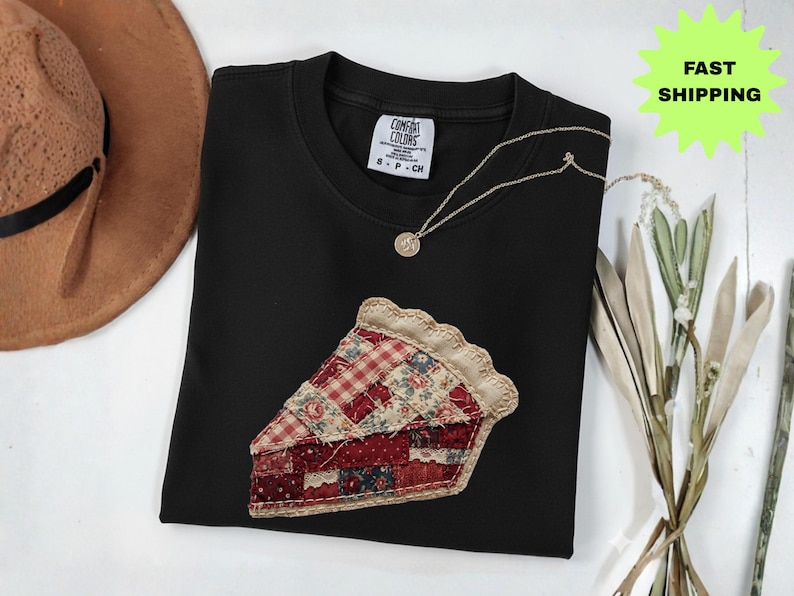 May include: A black crew neck sweatshirt with a pie slice applique. The pie slice is made of various patterned fabrics in red, cream, and blue. A gold necklace with a pendant is on the shirt. A brown hat is in the background.