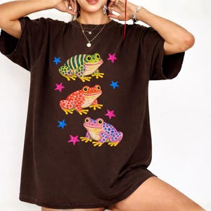 May include: A dark brown t-shirt with three frog illustrations in green, red, and purple. Each frog has a unique pattern, with pink and blue star shapes around them. The t-shirt has a loose, relaxed fit.
