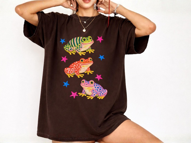 May include: A dark brown t-shirt with three frog illustrations in green, red, and purple. Each frog has a unique pattern, with pink and blue star shapes around them. The t-shirt has a loose, relaxed fit.