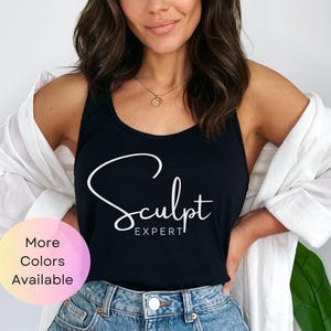 Pode incluir: Um top preto de alças com o texto "Sculpt Expert" em letras brancas. A modelo está usando o top com uma camisa branca por cima e jeans azul.