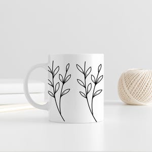 May include: White ceramic mug with a black and white botanical design of two sprigs with leaves. A ball of twine is to the right of the mug. The mug is on a white surface.