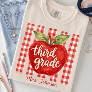May include: A cream-colored t-shirt featuring a red and white gingham pattern background with a large red apple graphic. The apple has green leaves and the text third grade written in white script. Below the apple is the text Mrs. Johnson.