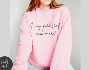 May include: A light pink sweatshirt with the text "In my published author era" in black script. The sweatshirt is a solid color and has long sleeves. The text includes a small heart.