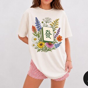 May include: An off-white t-shirt featuring a central graphic of a green mahjong tile with the Chinese character for 'Fa' surrounded by a colourful arrangement of wildflowers including blue, orange, yellow, and purple blossoms with green leaves.