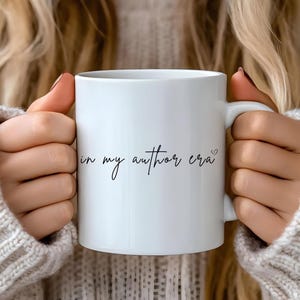 May include: White ceramic mug with black text that reads "in my author era" with a small heart symbol.