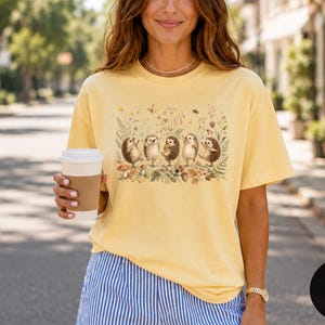 Puede incluir: Una camiseta de manga corta de color amarillo p&aacute;lido con un estampado gr&aacute;fico de cinco erizos de pie en fila entre varias flores silvestres, helechos y peque&ntilde;os insectos. Los erizos est&aacute;n representados en tonos marr&oacute;n claro con texturas.