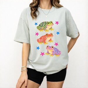 May include: A pale green t-shirt with a design of three cartoon frogs in vibrant colours. The frogs are green, red, and purple, with blue and pink star accents. The shirt has a relaxed, oversized style.