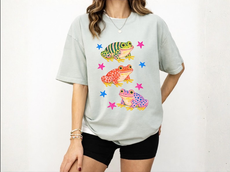 May include: A pale green t-shirt with a design of three cartoon frogs in vibrant colours. The frogs are green, red, and purple, with blue and pink star accents. The shirt has a relaxed, oversized style.
