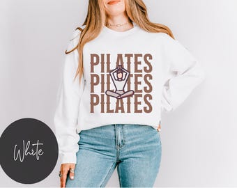 May include: White sweatshirt with the text "PILATES" repeated three times in brown text. A stylised image of a person doing a yoga pose is in the centre of the text.