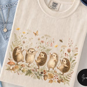 Puede incluir: Una camiseta color marfil con un estampado gr&aacute;fico de cinco erizos bailando entre flores silvestres, helechos, setas y bellotas. Una peque&ntilde;a mariposa y una abeja aparecen sobre los erizos. La camiseta tiene una etiqueta Comfort Colors en el cuello.