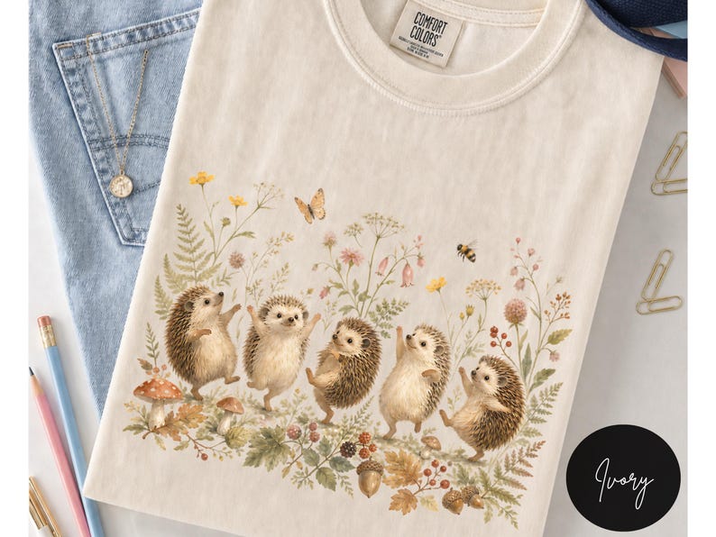 Puede incluir: Una camiseta color marfil con un estampado gr&aacute;fico de cinco erizos bailando entre flores silvestres, helechos, setas y bellotas. Una peque&ntilde;a mariposa y una abeja aparecen sobre los erizos. La camiseta tiene una etiqueta Comfort Colors en el cuello.