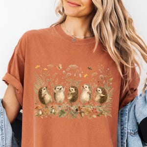 Puede incluir: Una camiseta de manga corta color &oacute;xido con un estampado gr&aacute;fico de cinco erizos de pie en fila en medio de una escena bot&aacute;nica detallada con flores silvestres, helechos, setas, una mariposa y una abeja en tonos tierra y apagados.