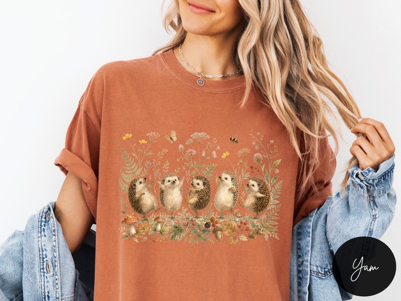 Puede incluir: Una camiseta de manga corta color &oacute;xido con un estampado gr&aacute;fico de cinco erizos de pie en fila en medio de una escena bot&aacute;nica detallada con flores silvestres, helechos, setas, una mariposa y una abeja en tonos tierra y apagados.