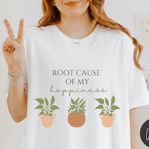 May include: White t-shirt with the text "Root Cause of My Happiness" and three potted plants with green leaves.