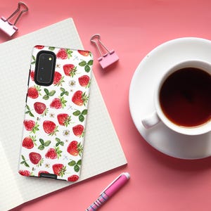 May include: A white phone case featuring a pattern of red strawberries and green leaves. The case has a black camera cutout and a black border.
