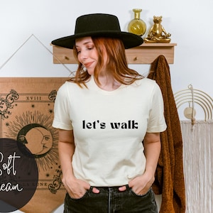 May include: A woman wearing a white t-shirt with the text "let's walk" in black lettering. She is wearing a black hat and jeans.