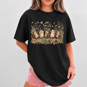 Puede incluir: Una camiseta negra de manga corta con un estampado gr&aacute;fico de cinco erizos de pie en fila entre varias flores silvestres, helechos, setas y una peque&ntilde;a mariposa. Los erizos est&aacute;n representados en un color marr&oacute;n claro con texturas detalladas.