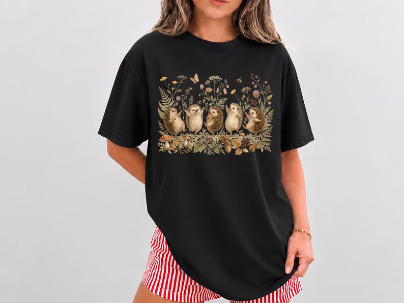 Puede incluir: Una camiseta negra de manga corta con un estampado gr&aacute;fico de cinco erizos de pie en fila entre varias flores silvestres, helechos, setas y una peque&ntilde;a mariposa. Los erizos est&aacute;n representados en un color marr&oacute;n claro con texturas detalladas.