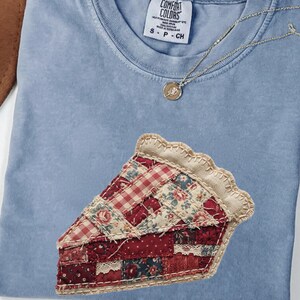 May include: A pale blue Comfort Colors t-shirt featuring a pie slice applique. The pie slice is made of various red and floral patterned fabrics. A gold necklace with a pendant is on the shirt.