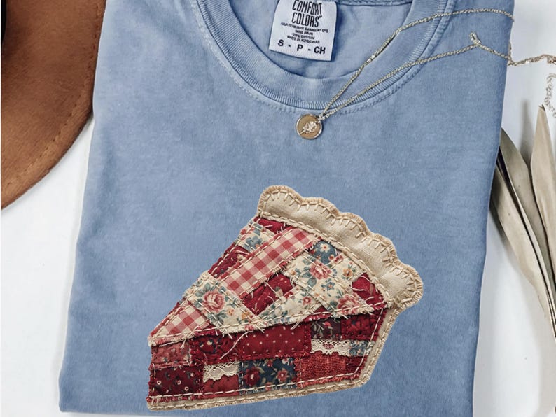 May include: A pale blue Comfort Colors t-shirt featuring a pie slice applique. The pie slice is made of various red and floral patterned fabrics. A gold necklace with a pendant is on the shirt.