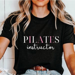 Pode incluir: Uma camiseta preta com o texto "PILATES instructor" em branco com o "I" em rosa.