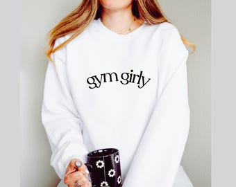 May include: White sweatshirt with the text "gym girly" in a black, stylish font. The person in the image is holding a black mug with white flower designs. The sweatshirt is a casual, comfortable style.