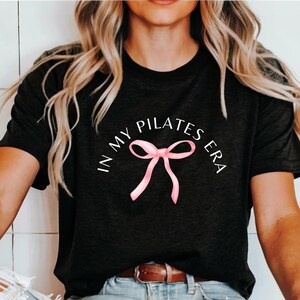 May include: A black t-shirt with the text "In My Pilates Era" in white lettering and a pink ribbon bow.