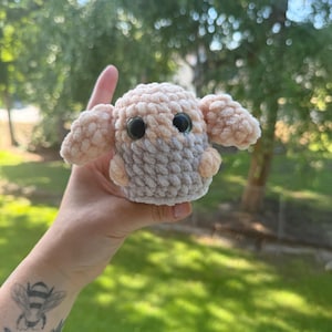 May include: A handmade crochet plushie in the shape of a small animal, featuring a light beige and gray color scheme. The plushie has large black eyes and small, rounded ears. The soft texture and rounded shape suggest a cuddly toy.
