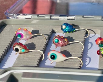 May include: A gray tackle box with several colorful fishing lures. The lures have large, detailed eyes and are various colors, including red, blue, and pink, with glitter accents. Each lure has a curved hook.