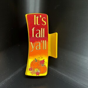 May include: Yellow and red hair clip with the text "It's fall y'all" in gold lettering. The clip features a gradient of colors and a pumpkin illustration.