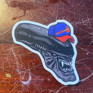 Op de afbeelding: Een zwart en grijs alienhoofd met een blauwe en rode baseballpet met de tekst "MINX" op de voorkant.