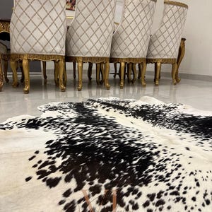 May include: A black and white cowhide rug with a white background and black spots. The rug is in front of a set of gold-trimmed chairs with white upholstery.