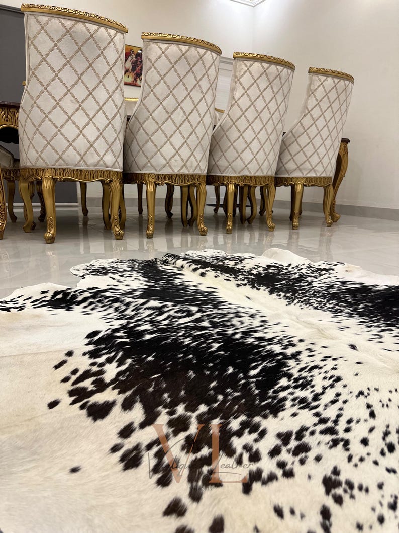 May include: A black and white cowhide rug with a white background and black spots. The rug is in front of a set of gold-trimmed chairs with white upholstery.