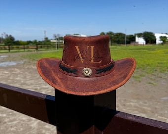 May include: A brown leather cowboy hat with a black band and a silver buckle. The hat has the text "Vintage Leather" embossed on the crown.