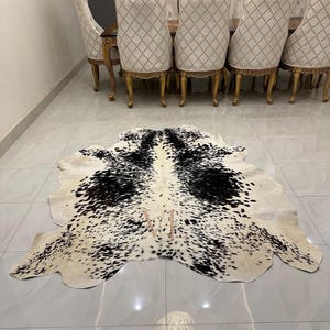 May include: A black and white cowhide rug with a spotted pattern. The rug is laid out on a white tiled floor in front of a set of gold dining chairs.