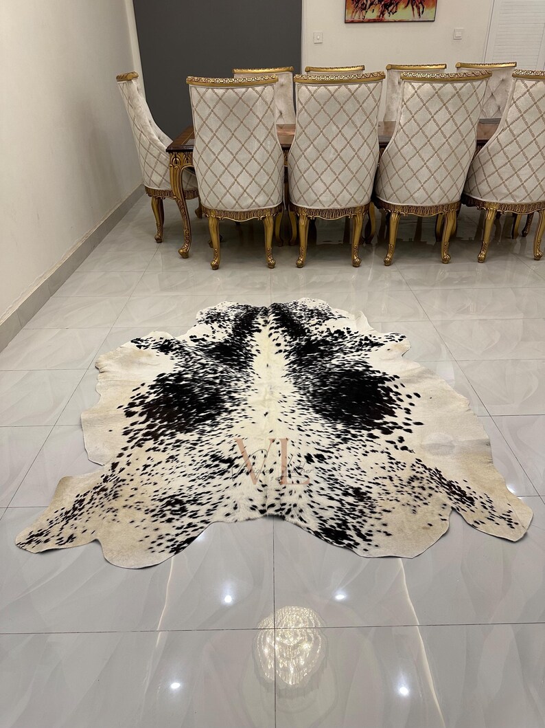 May include: A black and white cowhide rug with a spotted pattern. The rug is laid out on a white tiled floor in front of a set of gold dining chairs.