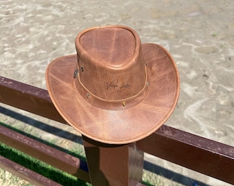 May include: A brown leather cowboy hat with a wide brim and a signature on the front. The hat is sitting on a wooden fence post.