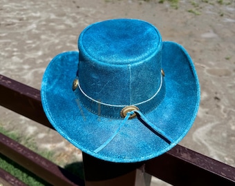 May include: A blue leather cowboy hat with a wide brim and a leather band with a decorative buckle. The hat is sitting on a wooden fence.