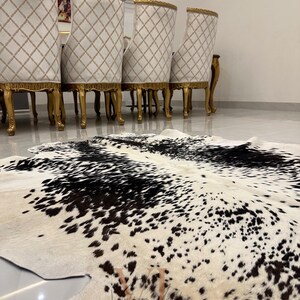 May include: A black and white cowhide rug with a spotted pattern. The rug is laid out on a white tiled floor in front of a set of gold-trimmed chairs.