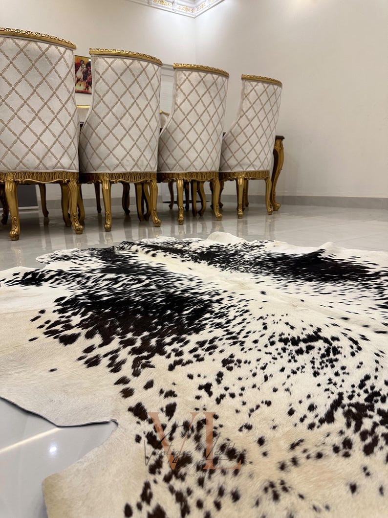 May include: A black and white cowhide rug with a spotted pattern. The rug is laid out on a white tiled floor in front of a set of gold-trimmed chairs.