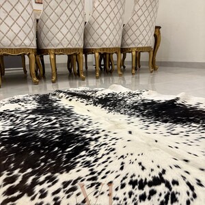 May include: A black and white cowhide rug with a natural, spotted pattern. The rug is laid out on a white tiled floor in front of a set of four gold-trimmed chairs.