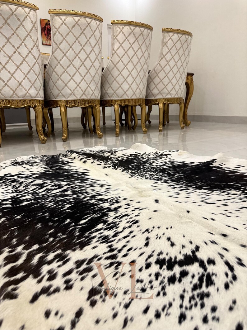 May include: A black and white cowhide rug with a natural, spotted pattern. The rug is laid out on a white tiled floor in front of a set of four gold-trimmed chairs.