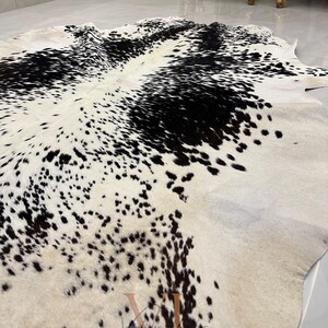 May include: A black and white cowhide rug with a spotted pattern. The rug is laid out on a light-colored floor.