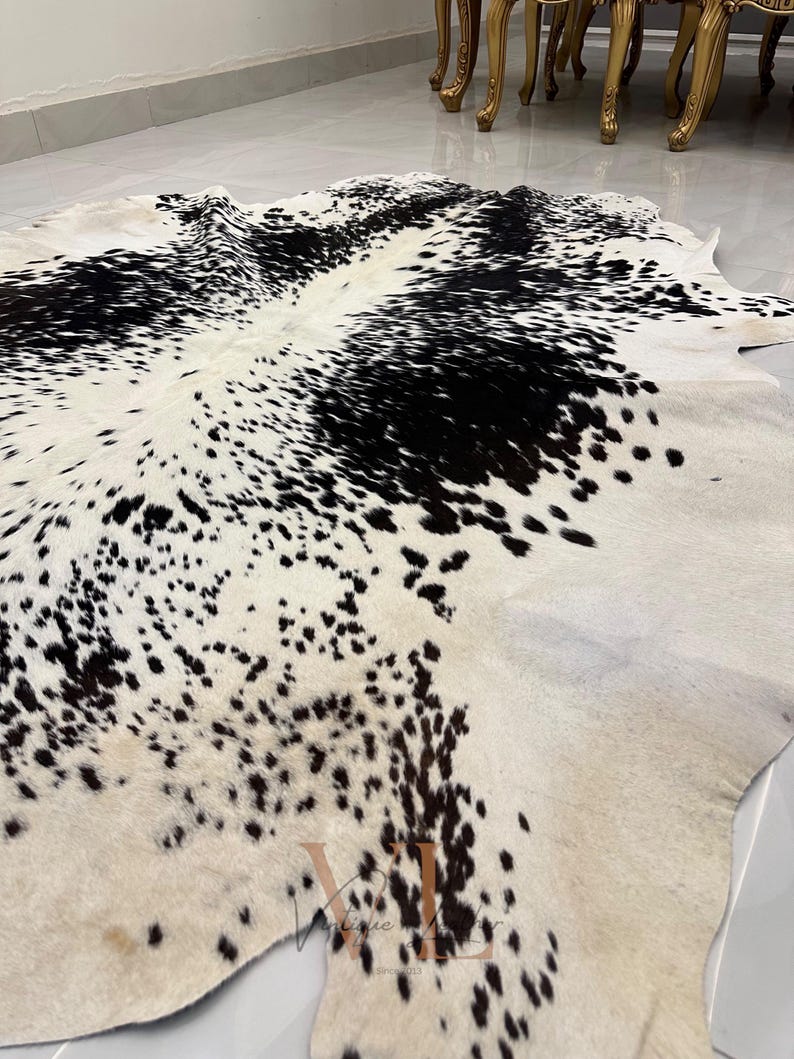 May include: A black and white cowhide rug with a spotted pattern. The rug is laid out on a light-colored floor.