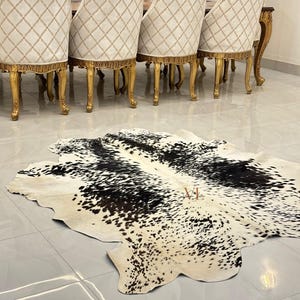 May include: A black and white cowhide rug with a natural, spotted pattern. The rug is laid out on a tiled floor in front of a set of gold-painted dining chairs.