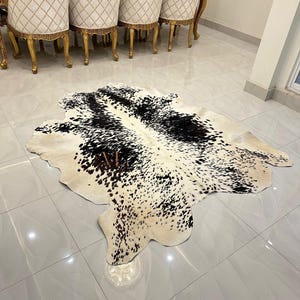 May include: A black and white cowhide rug with a natural, spotted pattern. The rug is laid out on a light grey tiled floor.